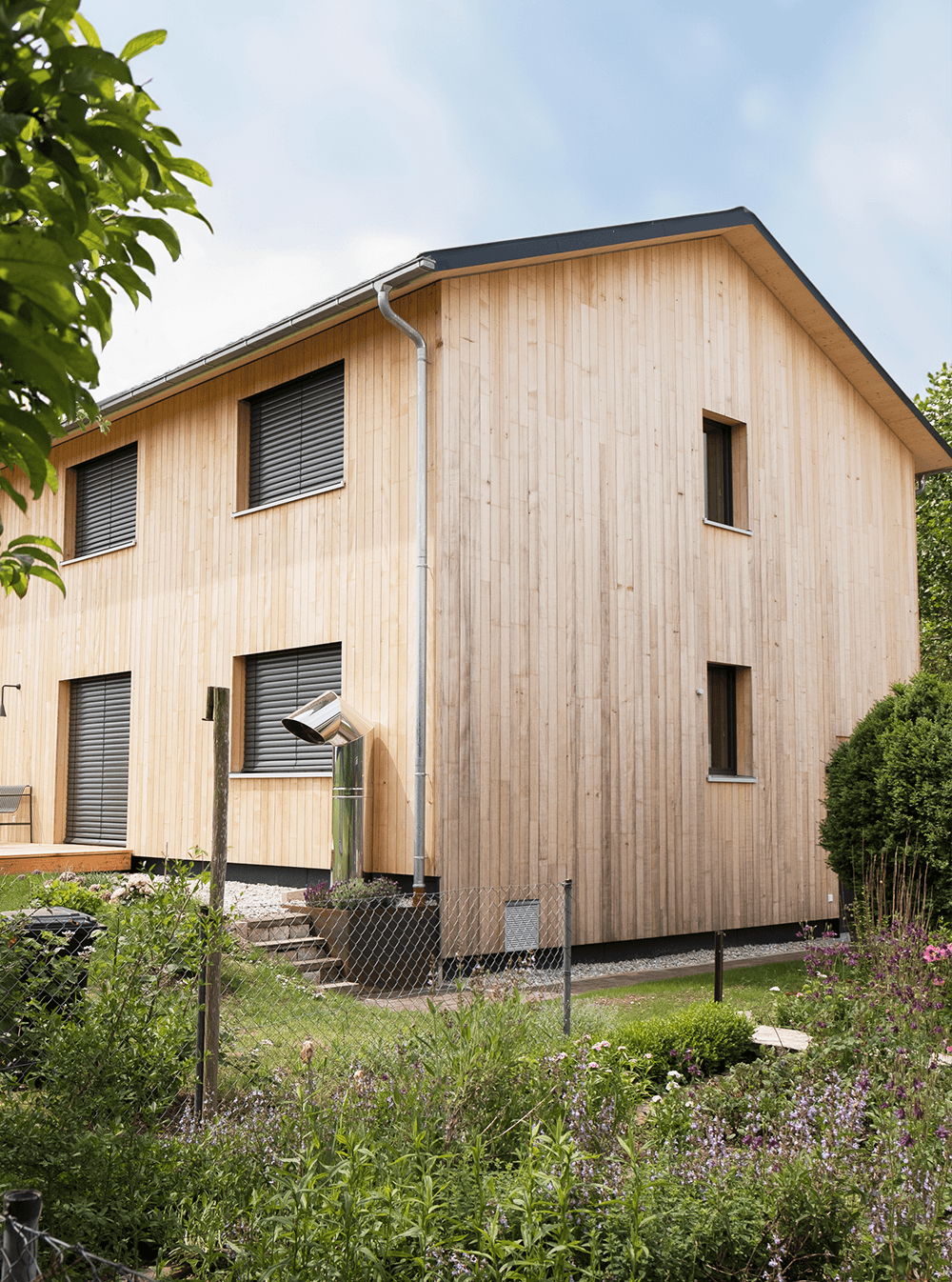 Zweistöckiges Holzhaus mit dunklen Jalousien und einem Schornstein; Grün im Vordergrund.