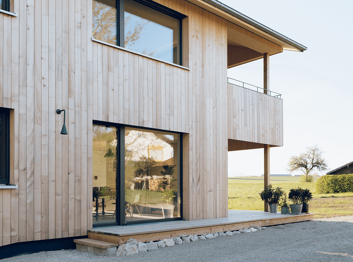 Modernes zweistöckiges Haus mit heller Holzverkleidung, großen Fenstern und einem Balkon mit Blick auf ein Feld.