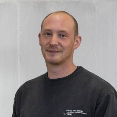 Ein Mann mit heller Haut und schütterem Haar, der ein schwarzes T-Shirt mit Rundhalsausschnitt trägt, lächelt.