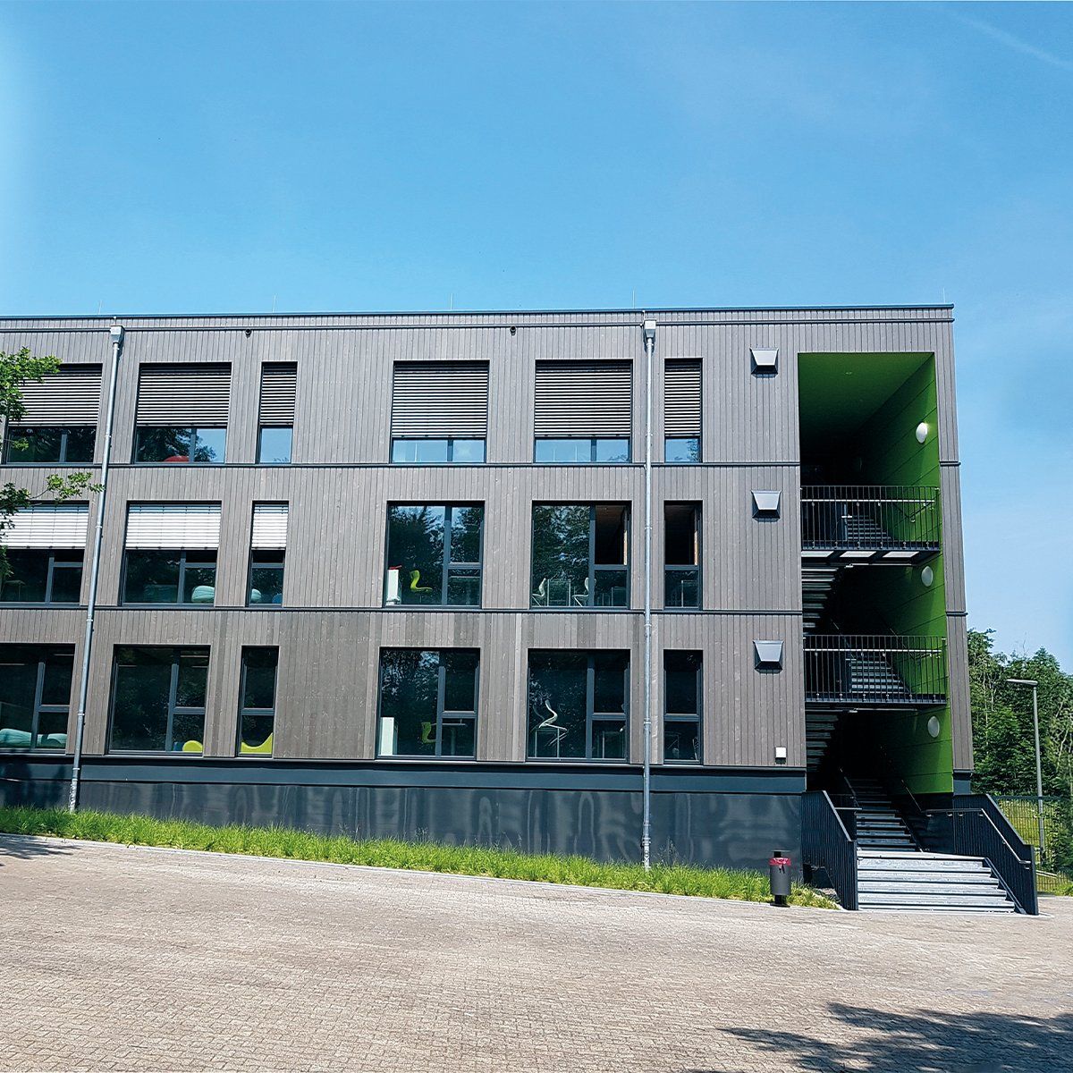 Modernes Mehrfamilienhaus mit Holzverkleidung, grünen Akzenten und Außentreppe unter blauem Himmel.