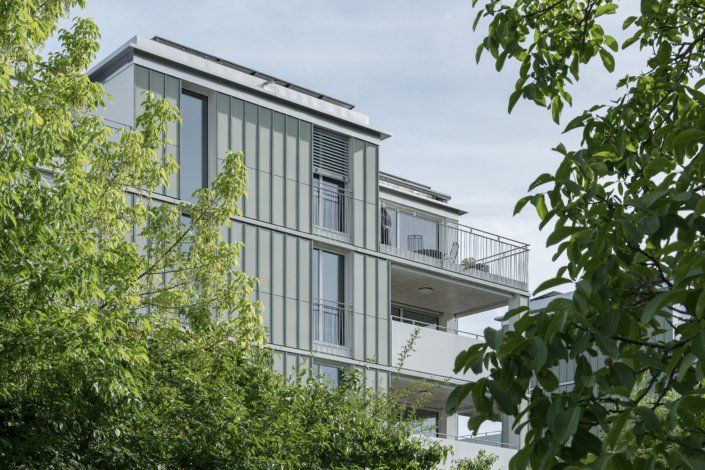 Modernes Gebäude mit Glaspaneelen, Balkonen und Flachdach, teilweise von grünen Bäumen verdeckt.