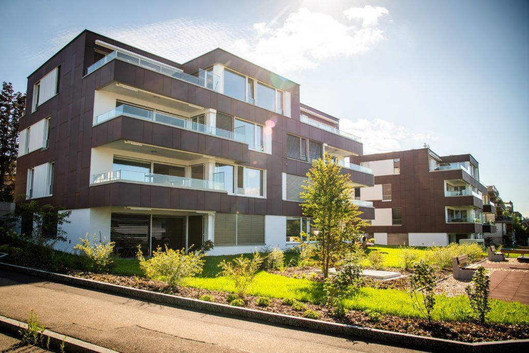 Modernes mehrstöckiges Wohnhaus mit dunkler Fassade, Glasbalkonen und Landschaftsgestaltung unter sonnigem Himmel.