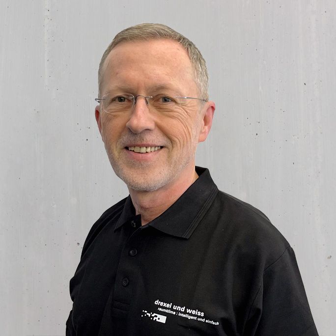 Mann mit Brille und kurzem Haar, der ein schwarzes Poloshirt trägt, lächelt vor einer hellen Wand.