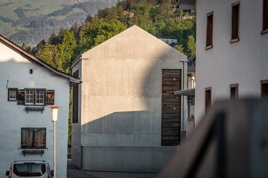 Graue Gebäudefassade zwischen zwei weißen Gebäuden. Holztür mit geschlossenen Fensterläden. Berge im Hintergrund.