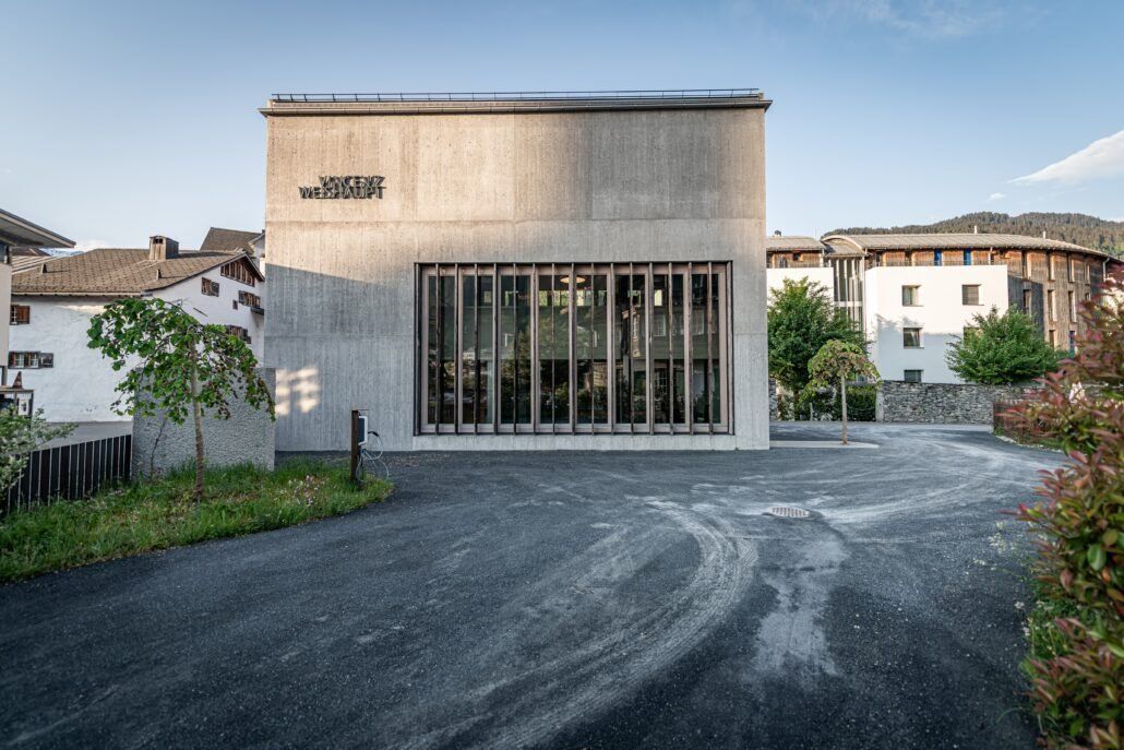 Modernes Betongebäude mit großem Fenster, Schotterparkplatz und umliegenden Bäumen in einem europäischen Dorf.