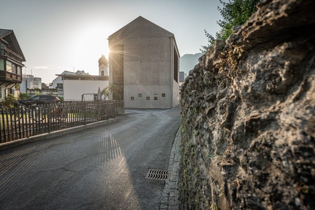 Eine Straße führt zu einem modernen Gebäude, dahinter helles Sonnenlicht, rechts davon eine Steinmauer.