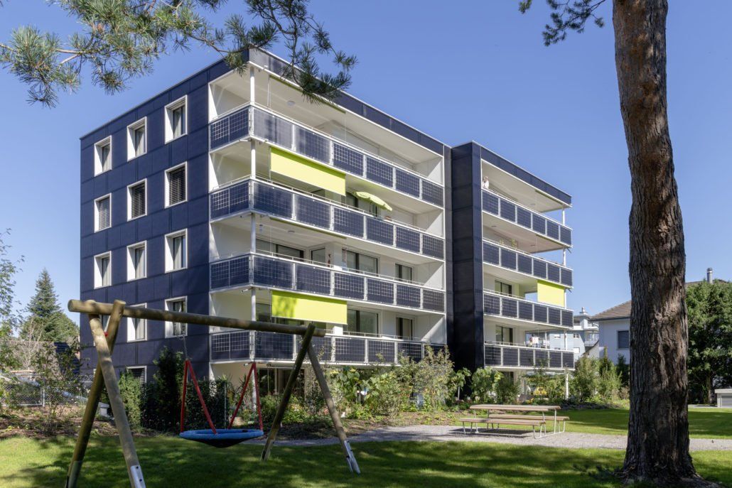 Modernes Mehrfamilienhaus mit Balkonen, Schaukel im Vordergrund, sonniger Tag.