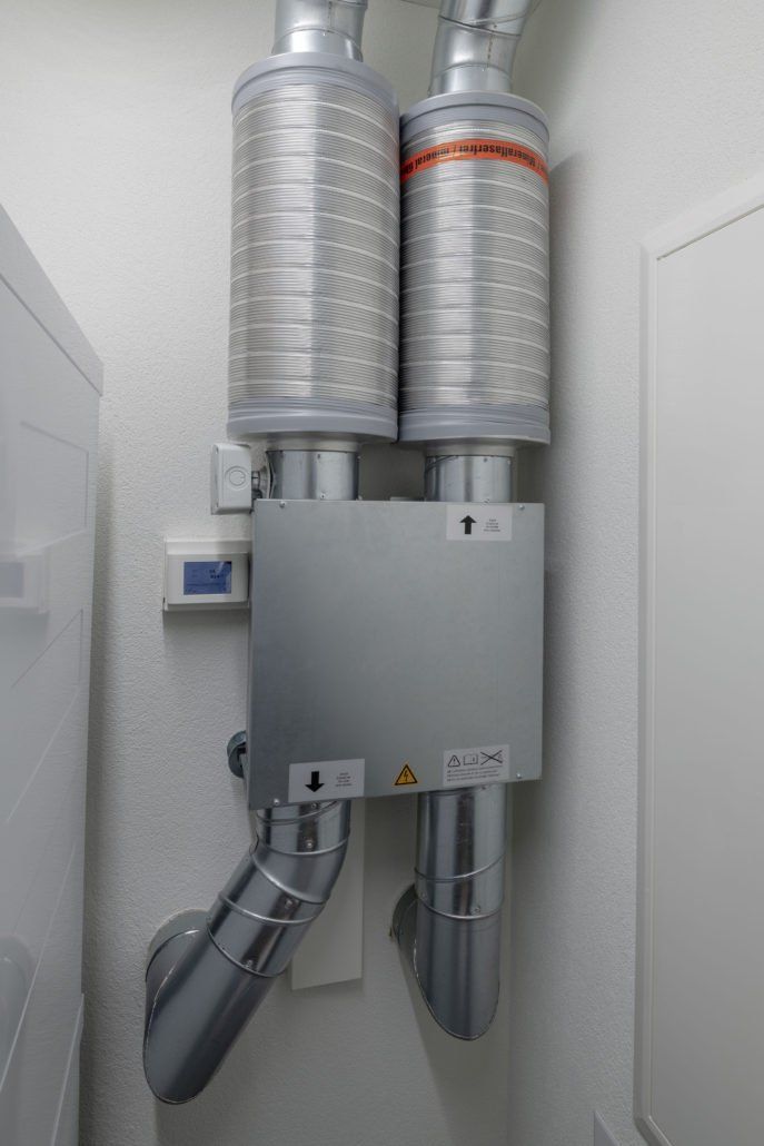 Heizungs-, Lüftungs- und Klimaanlage mit silbernen Luftkanälen und zylindrischen Filtern an einer weißen Wand.