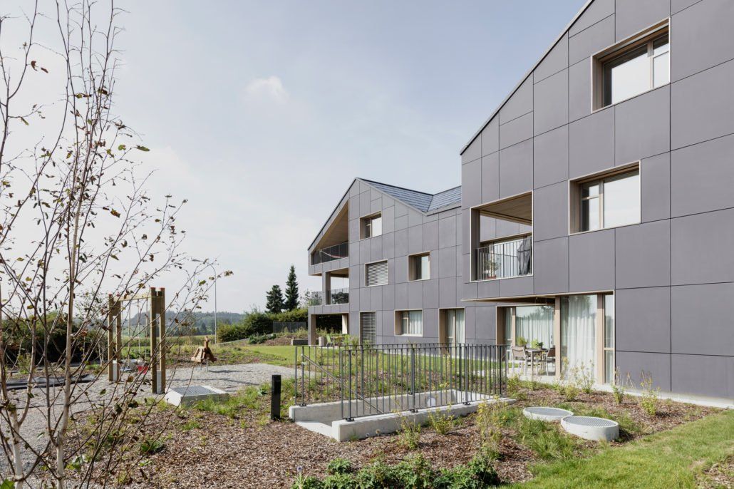 Modernes graues Gebäude mit Giebeldach und rechteckigen Fenstern, eingebettet in eine naturnahe Landschaft mit Gras und Pflanzen.