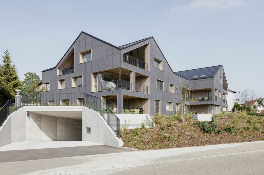Modernes graues Mehrfamilienhaus mit Holzelementen und Solarpaneelen, neben einer Garageneinfahrt aus Beton.