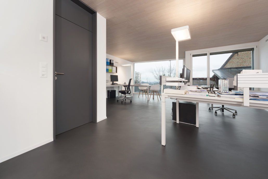 Modernes Bürointerieur mit dunklem Boden, grauer Tür und großen Fenstern. Schreibtisch und Stühle sind vorhanden.