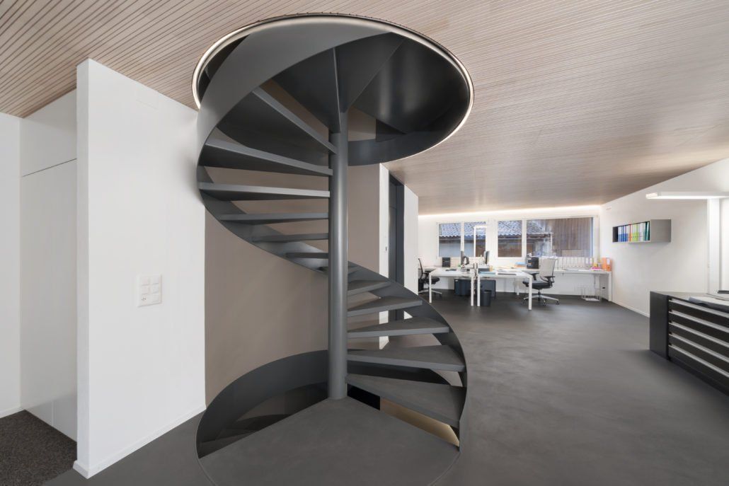Wendeltreppe in einem modernen Büro mit grauem Boden, weißen Wänden und Holzdecke.