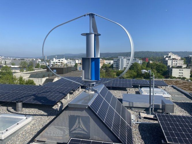 Windkraftanlage und Solarpaneele auf einem Gebäudedach vor blauem Himmel.
