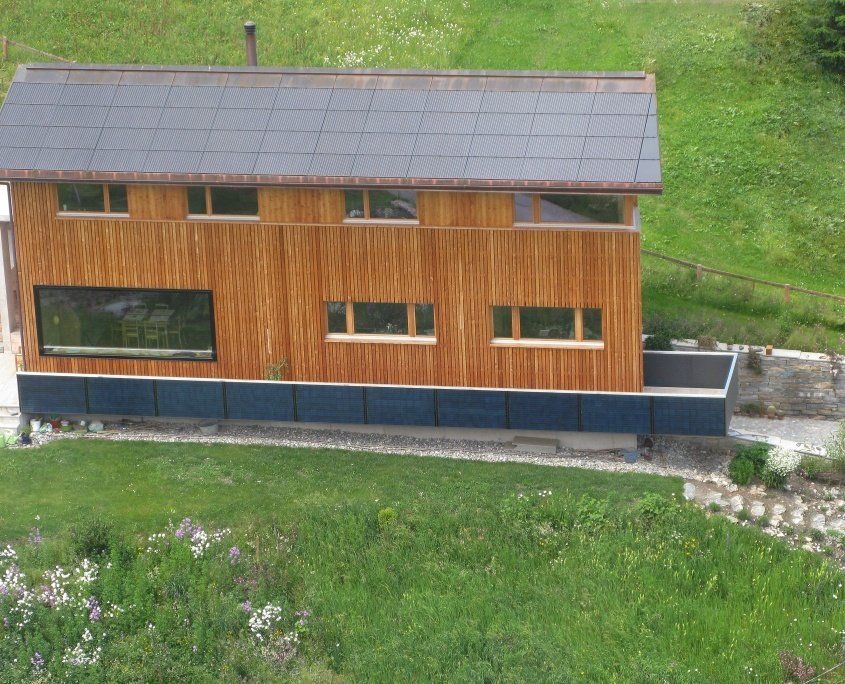 Holzhaus mit Solarpaneelen auf dem Dach, großem Fenster und grasbewachsener Umgebung.
