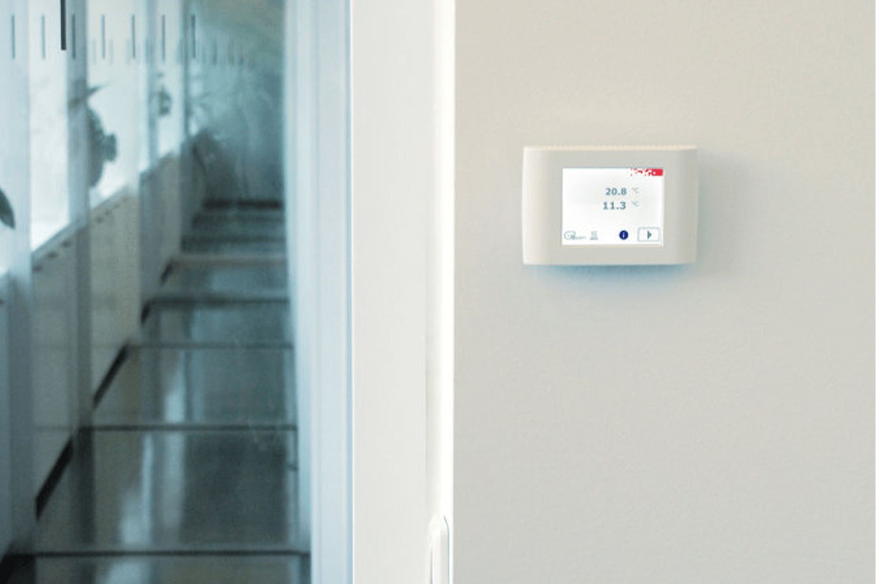 Weißer Thermostat an der Wand, der die Temperatur anzeigt; Flur mit Fenstern im Hintergrund.