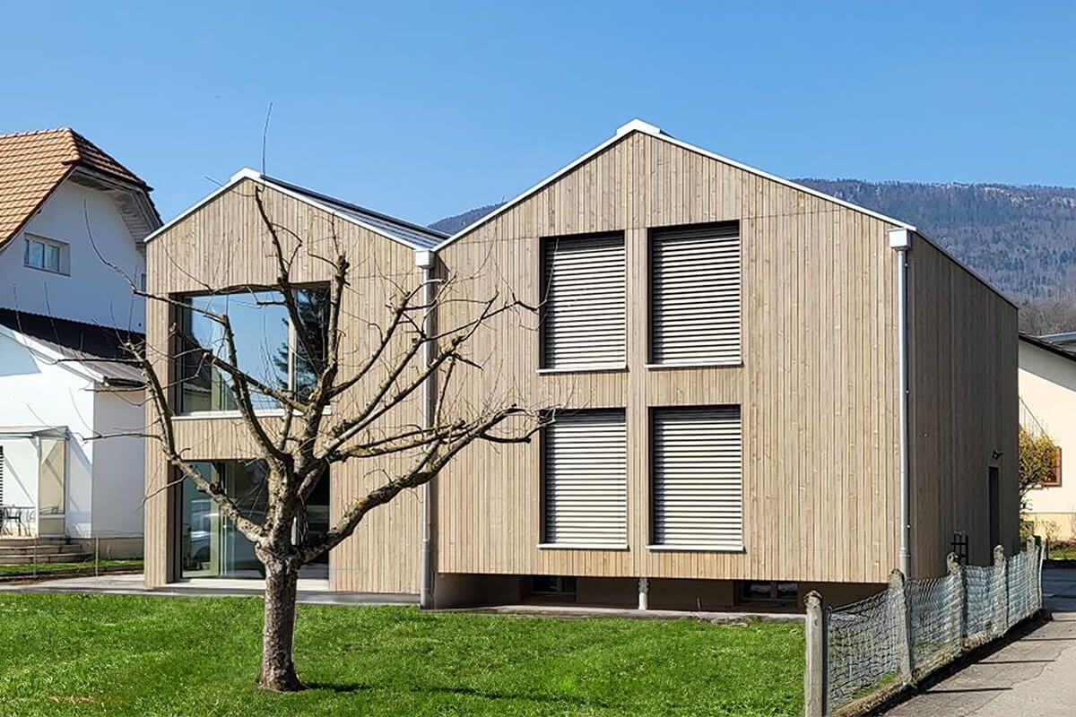 Modernes Holzhaus mit zwei Giebelseiten, großen Fenstern und Lamellenfensterläden auf einem grünen Rasen.