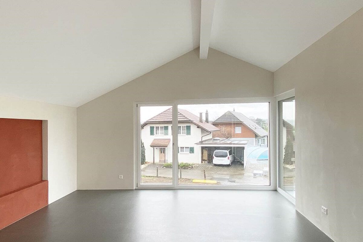Leerer, lichtdurchfluteter Raum mit großem Fenster und Blick auf Häuser. Weiße Wände, dunkler Boden und rote Wandverkleidung.