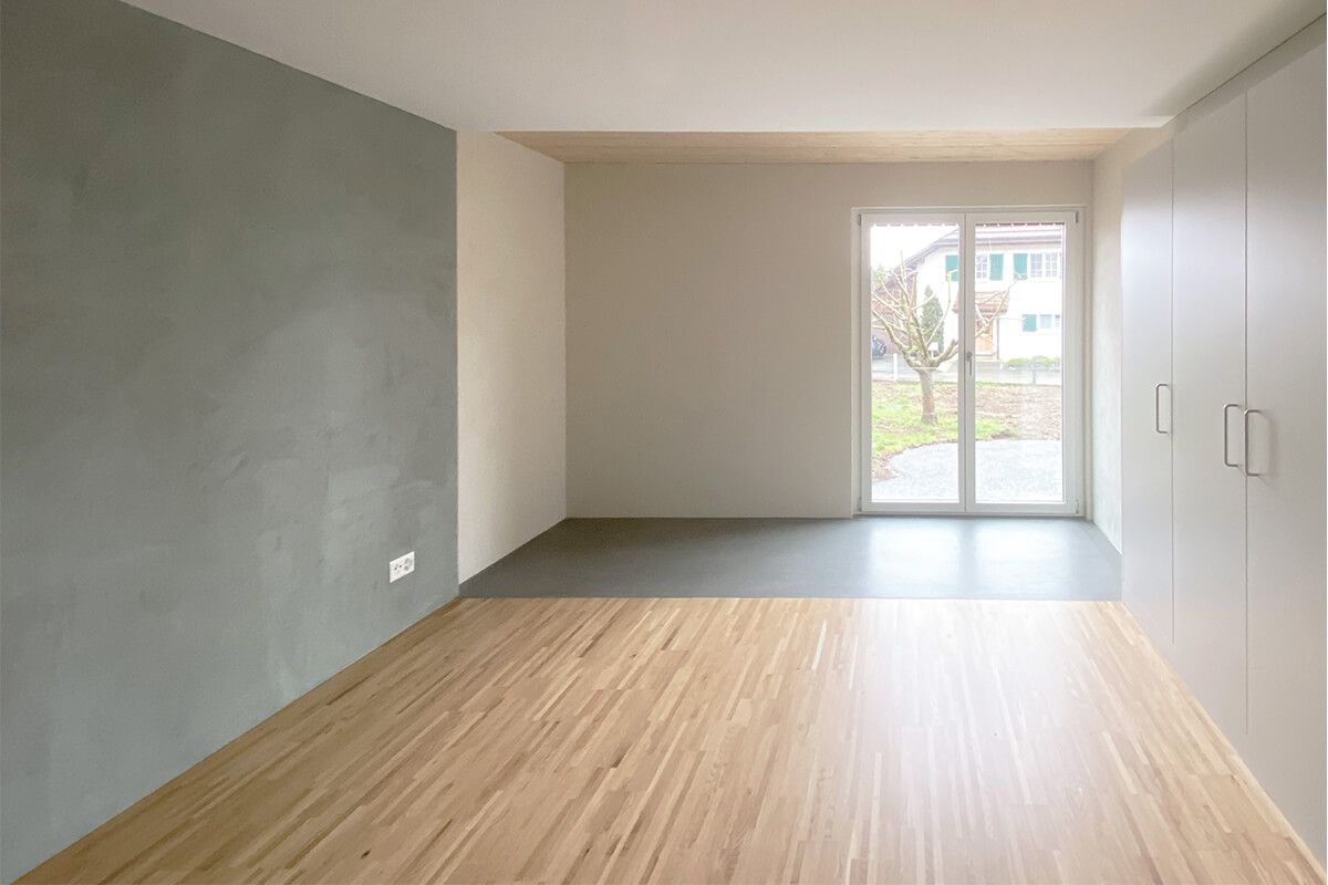 Leeres Zimmer mit Holzboden, einer grauen Akzentwand und einem großen Fenster.