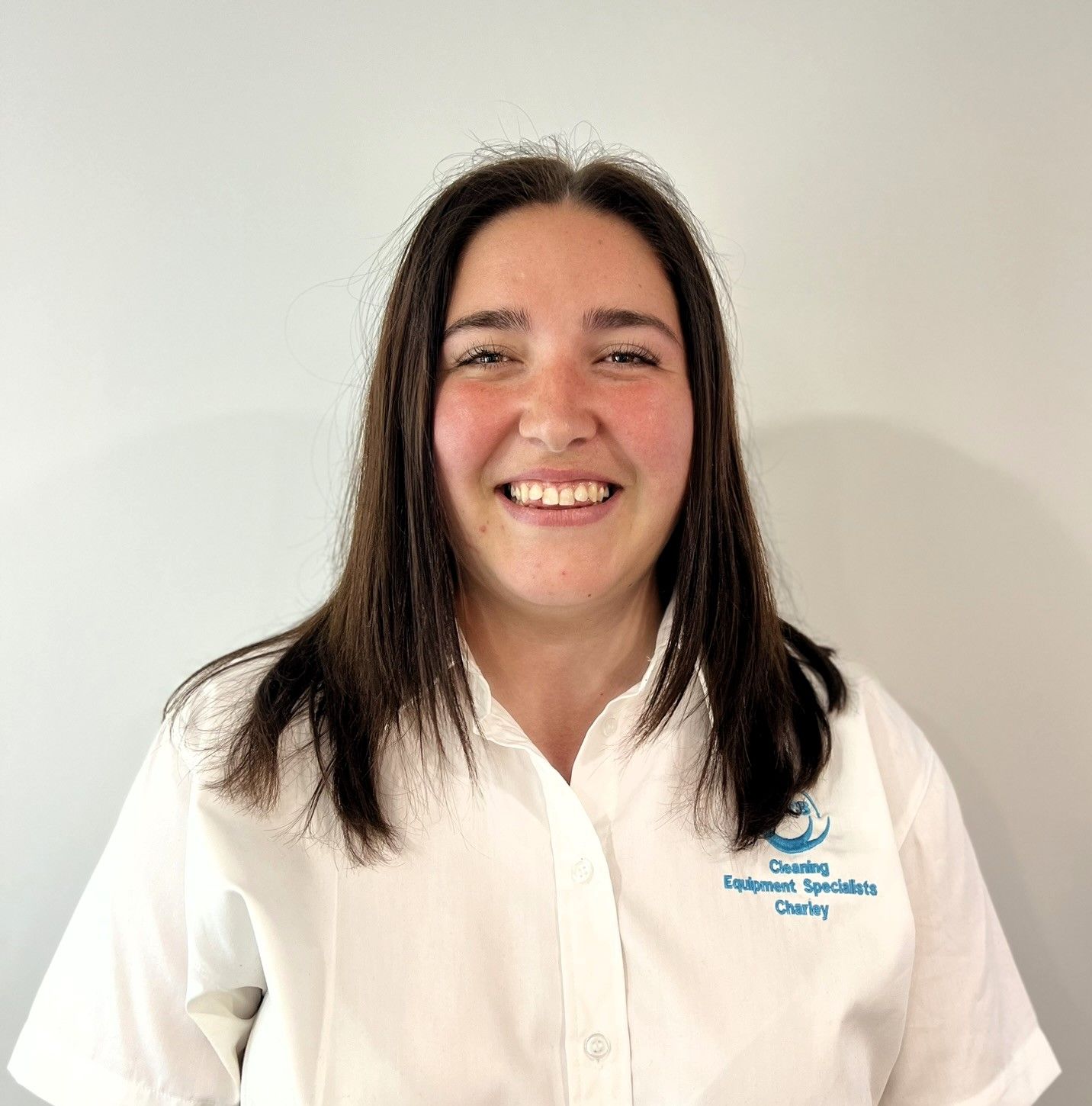 Woman wearing a white shirt with a logo, smiling in front of a white wall.