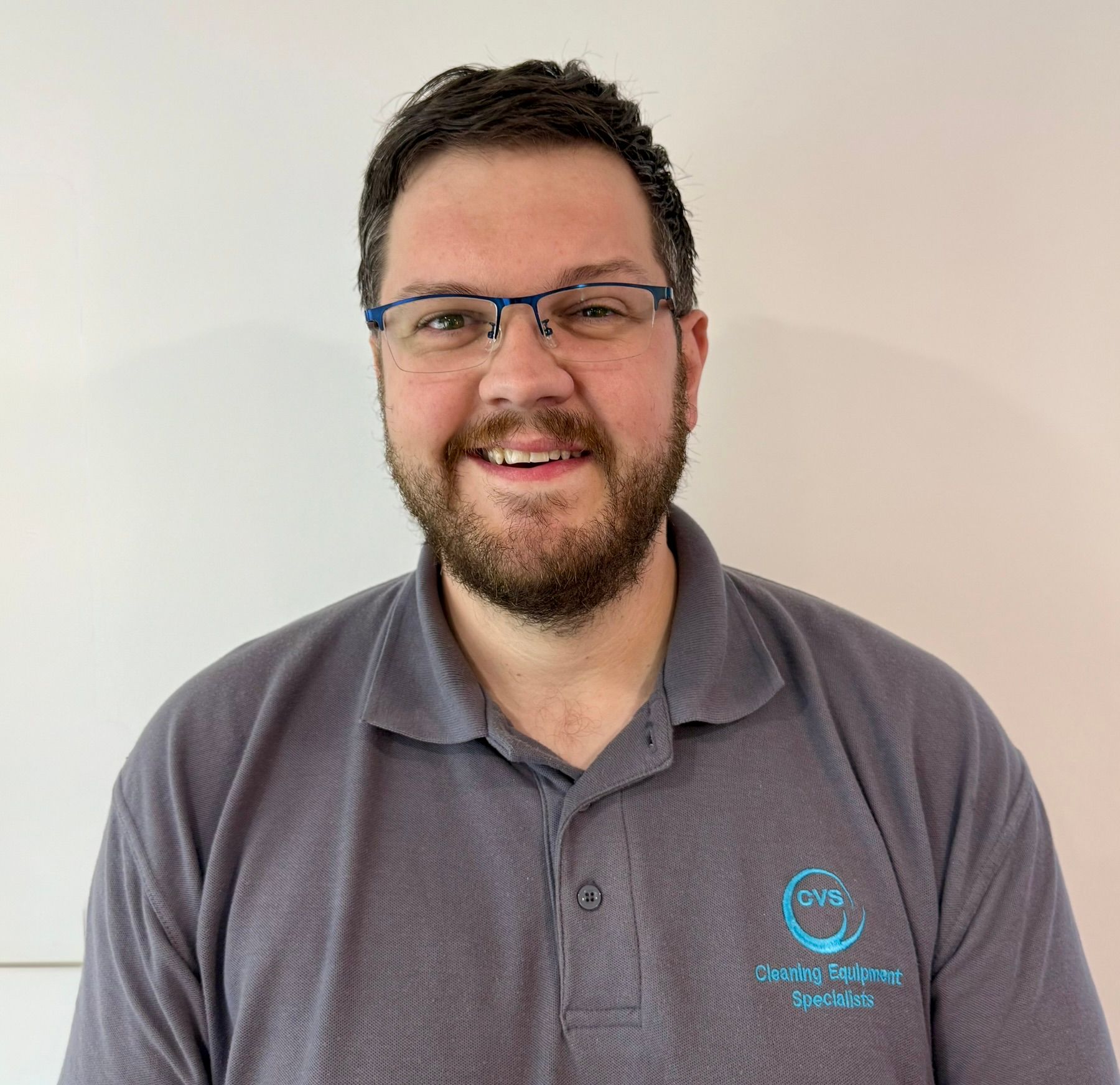 Man with glasses and a beard smiling, wearing a gray polo shirt with a logo, against a white background.