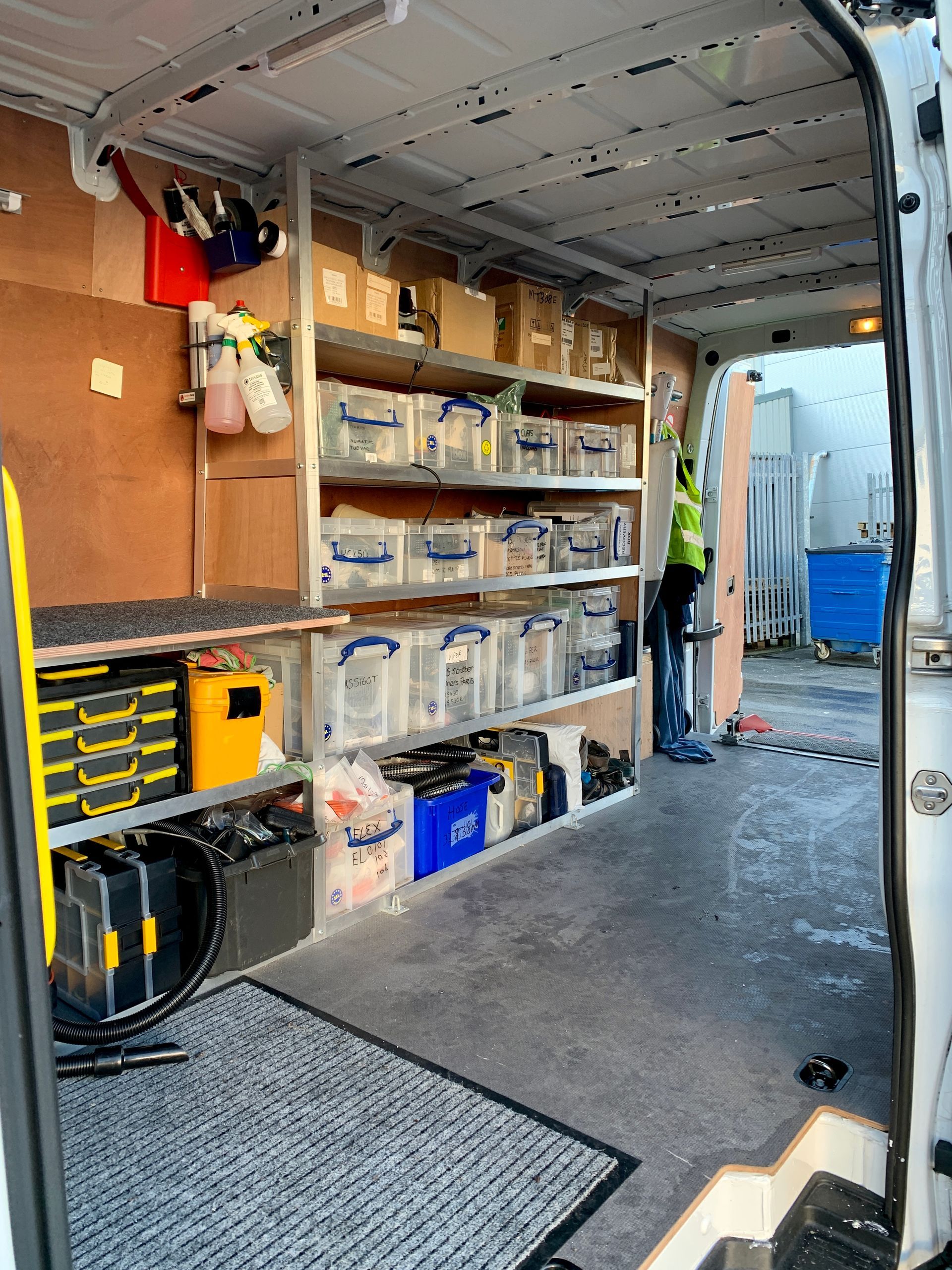 Interior of a cargo van filled with shelves, storage bins, and tools, a person standing by the open back doors.