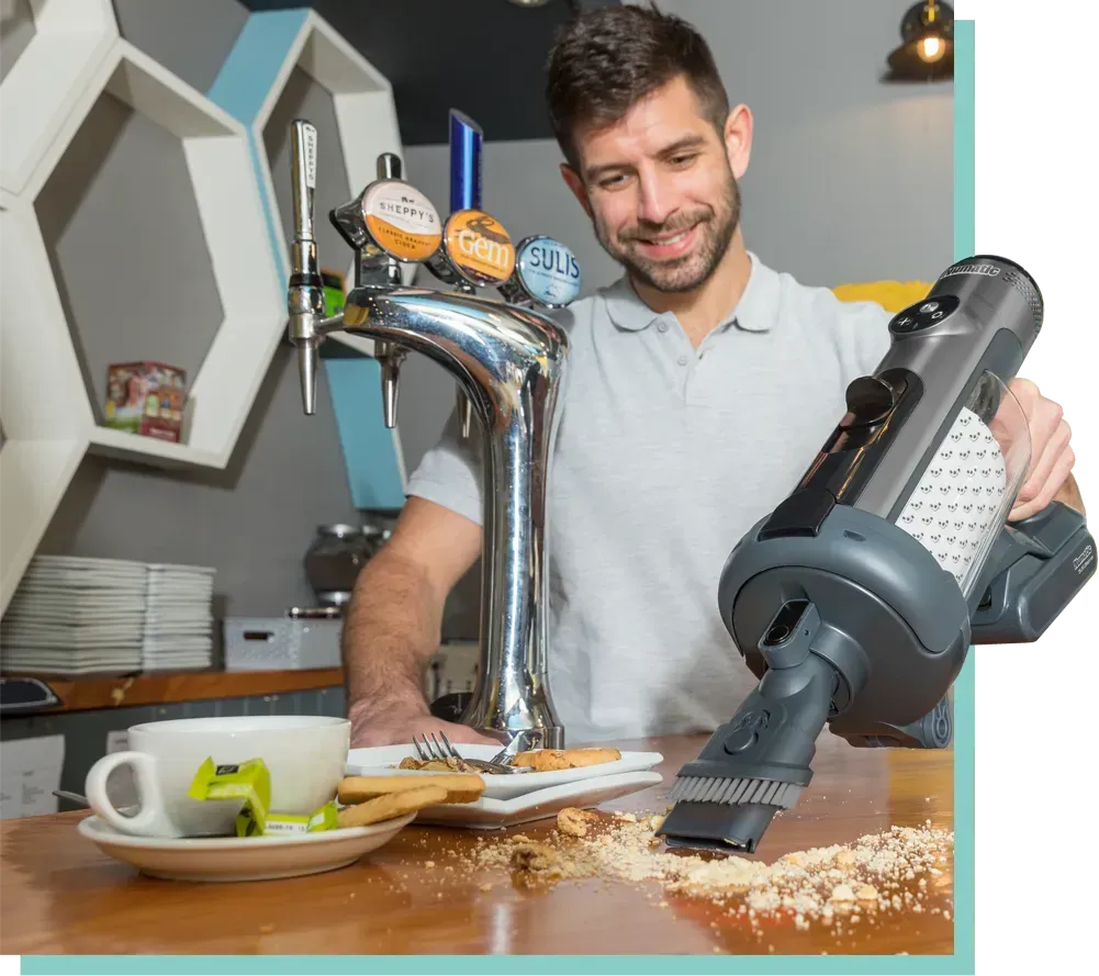 Man uses a handheld vacuum to clean a counter covered in crumbs in a bar.