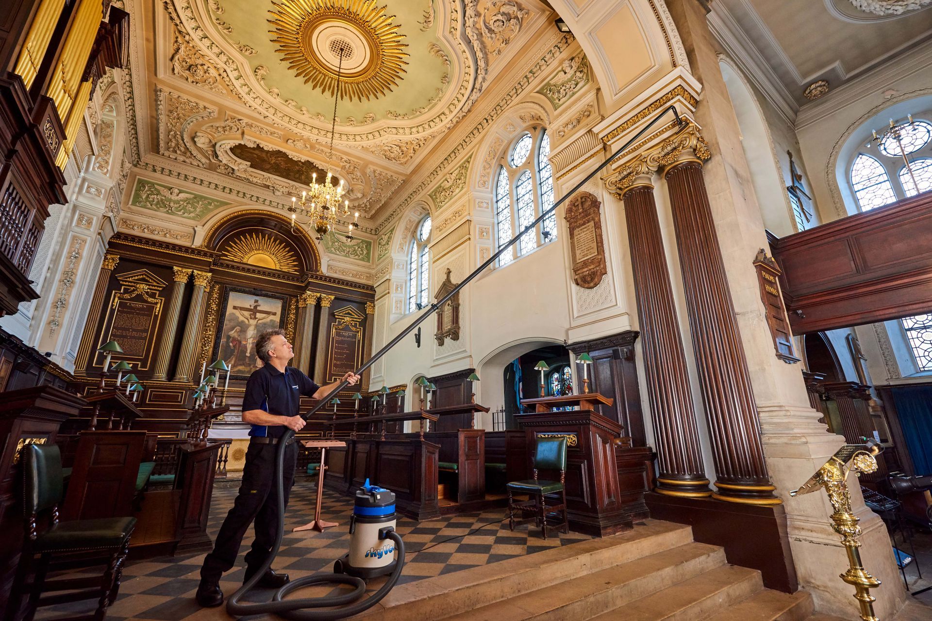 Person cleaning ornate church ceiling with long-handled vacuum.