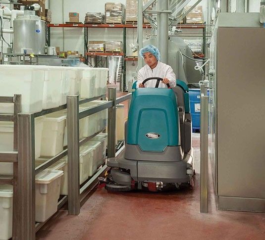 Teal and gray industrial floor scrubber with a seat and steering wheel.
