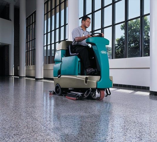 Person operating a teal floor cleaner machine in a building with large windows and a shiny floor.