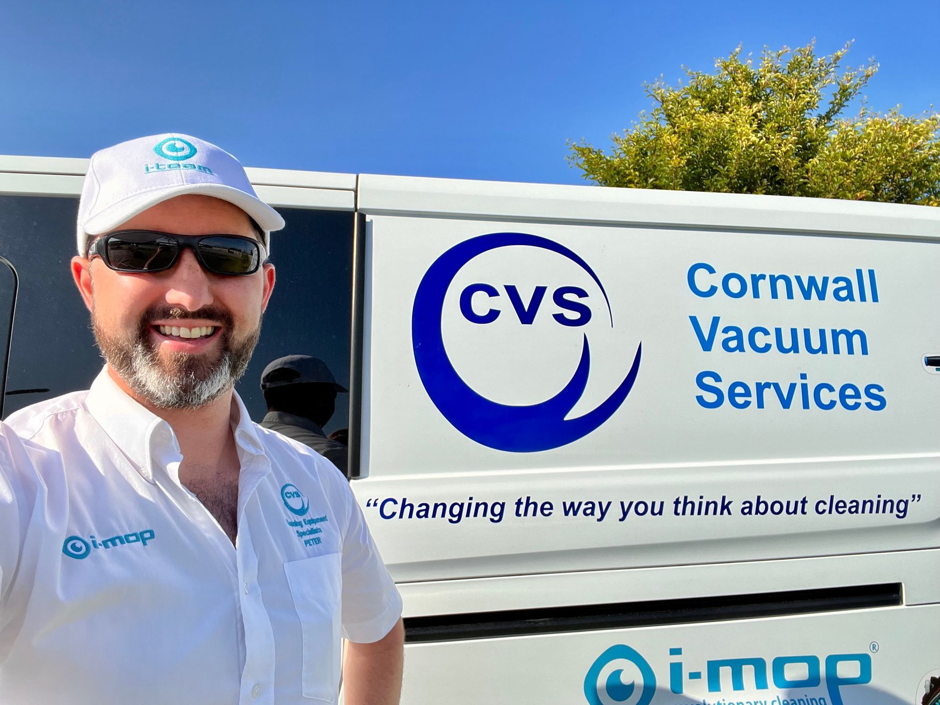 Man smiles in front of a Cornwall Vacuum Services van, wearing a hat, sunglasses, and a shirt with a logo.