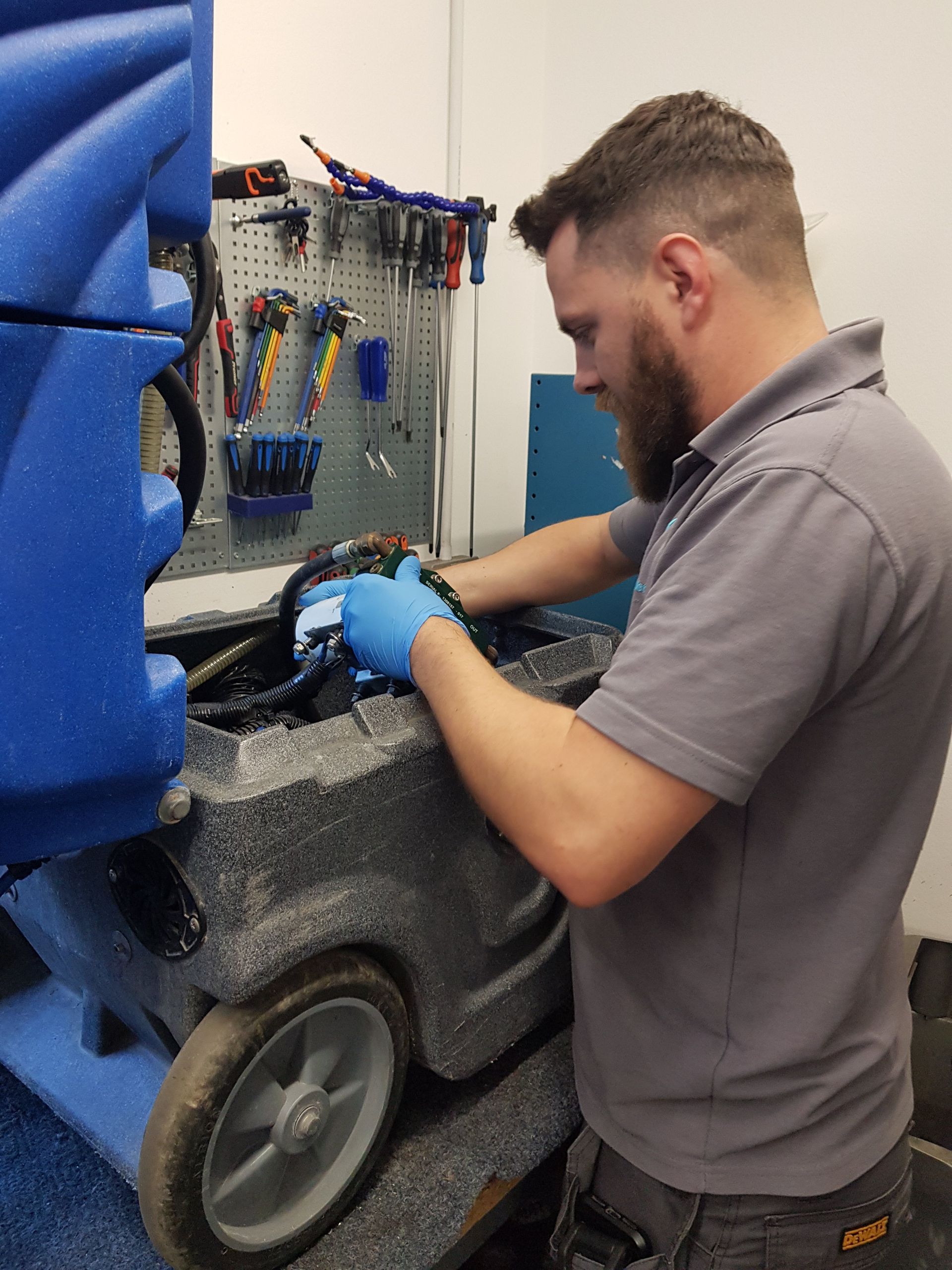 Mechanic with blue gloves works on a gray machine with a blue side; tools on a pegboard background.