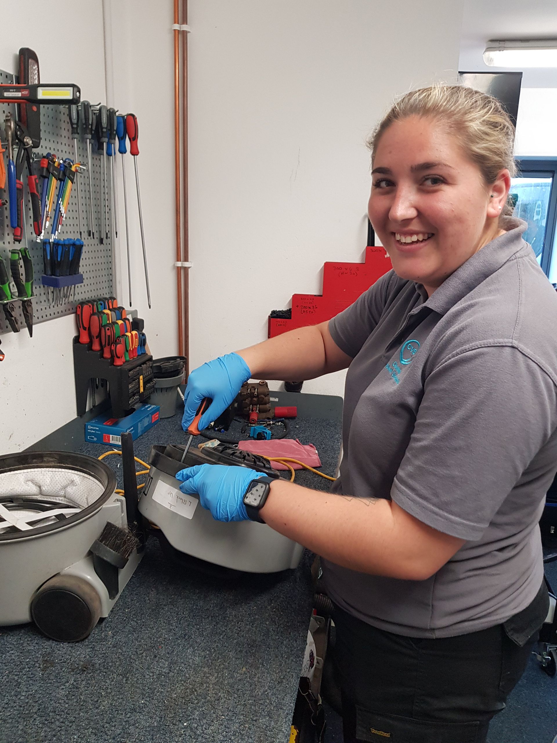 Woman in blue gloves smiles while working on machinery at a workbench with tools.