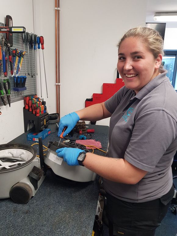 Woman in blue gloves smiles while working on machinery at a workbench with tools.