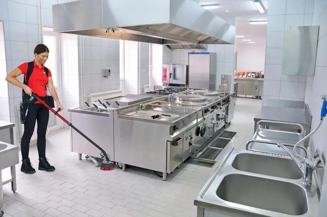 Person using a long-handled brush to clean a commercial kitchen floor, with stainless steel appliances and sinks.