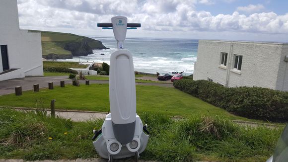 Robot on a grassy slope, with ocean view. Robot is white and gray, outdoors on a sunny day.
