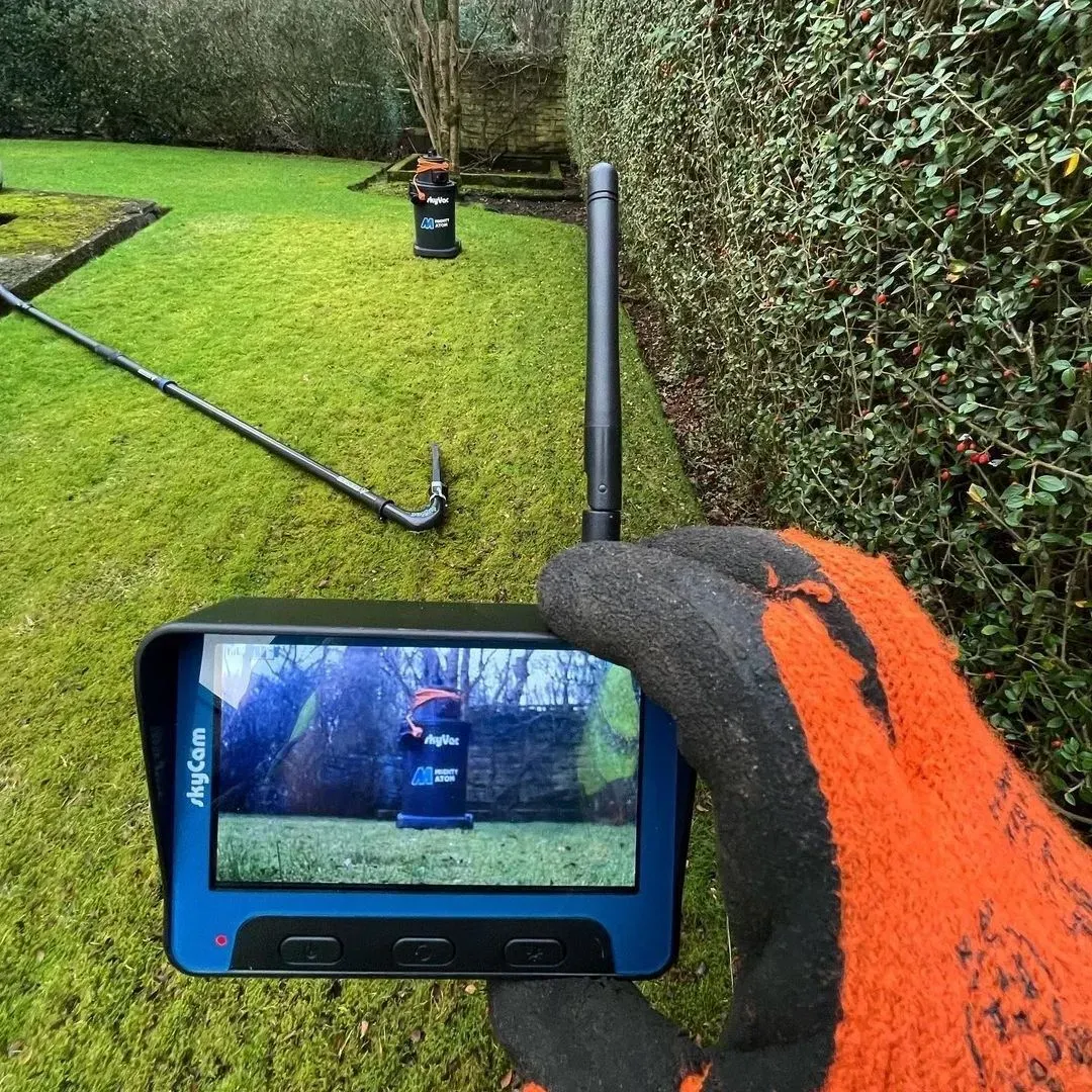 A gloved hand holds a monitor showing a camera view of a yard with equipment; lush green grass.