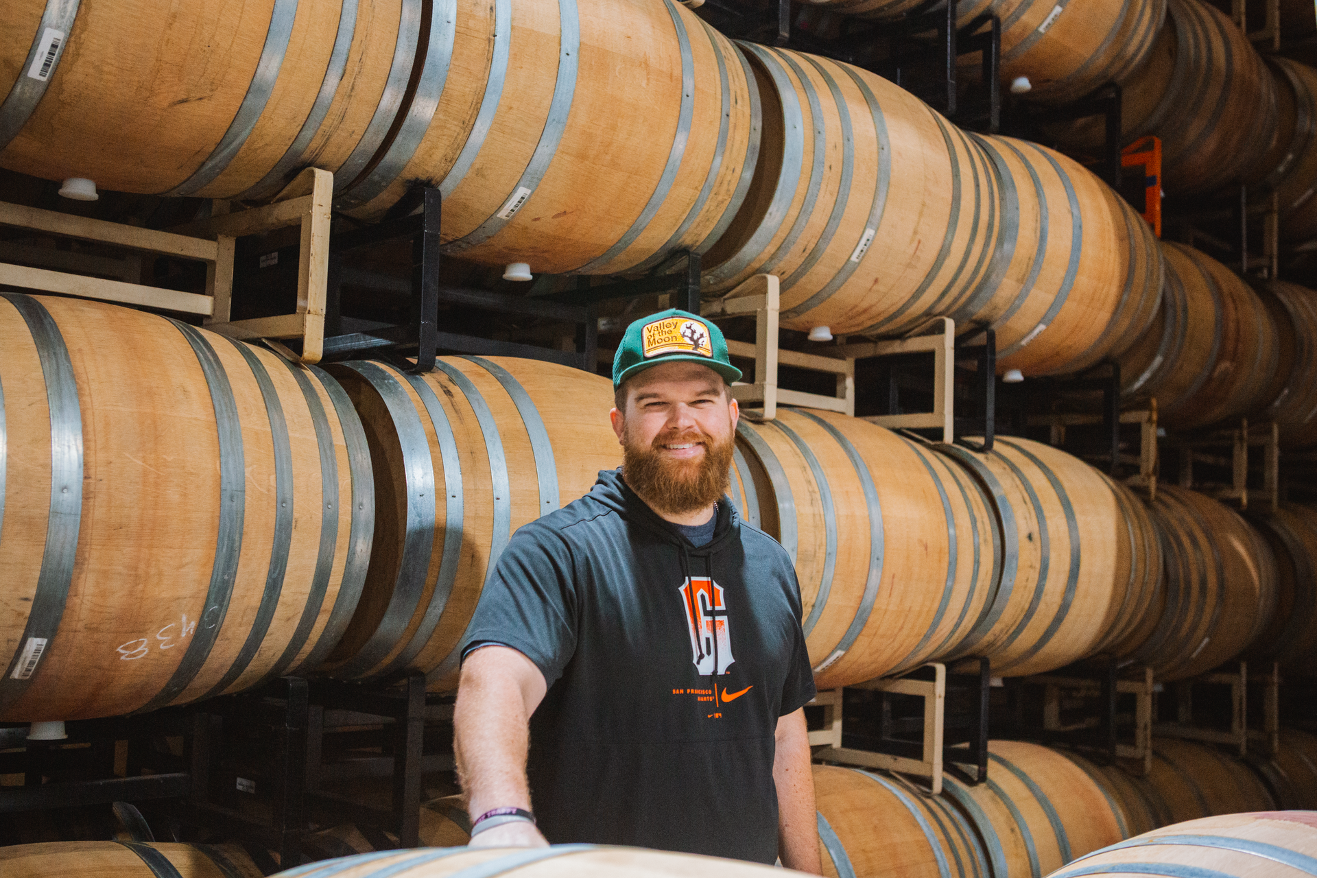 A man with a beard is standing in front of a row of wine barrels.