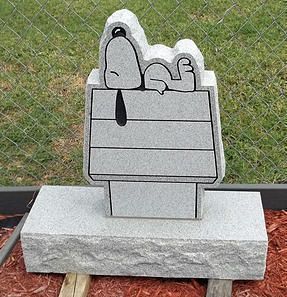 Cartoon-style gray stone sculpture of a face atop a pedestal, outdoors by a chain-link fence.