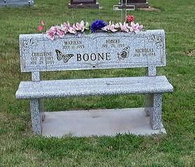 Memorial bench with engraved names and pink flowers on top, set in a grassy cemetery