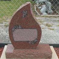 Red memorial stone with a blank plaque and floral engraving on a base outdoors by a fence.