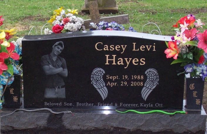 Black granite memorial gravestone with flowers, engraved “Casey Levi Hayes” and dates, in a grassy cemetery.