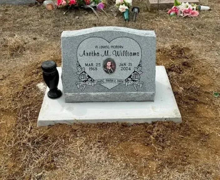 Gray gravestone with a photo and flowers on a bare dirt grave, next to a black vase.