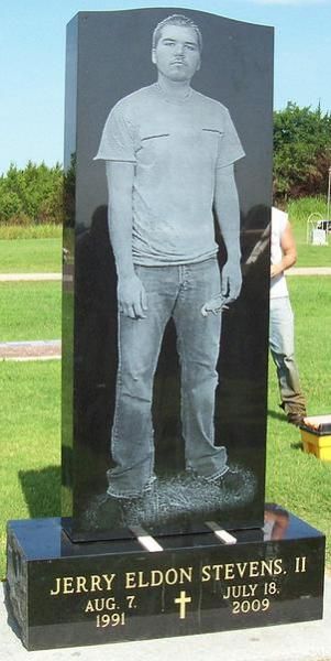 Gray stone memorial statue of Jerry Eldon Stevens II on a black pedestal outdoors with engraved dates.