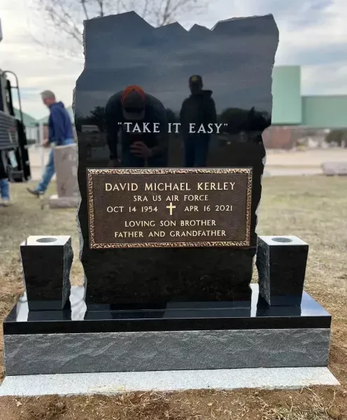 Black granite memorial headstone engraved “TAKE IT EASY” at a cemetery with flowers and a small vase below