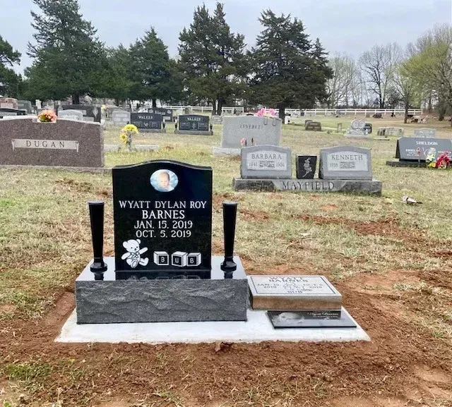 Black gravestone with flowers in a cemetery, surrounded by other headstones and trees.
