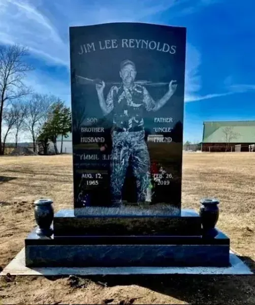 Memorial headstone for Jim Lee Reynolds in a grassy cemetery under a blue sky