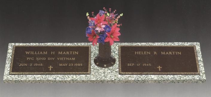Two bronze grave plaques with a vase of pink and purple flowers between them on gravel.