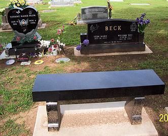 Black memorial bench in a cemetery with headstones and flowers behind it.