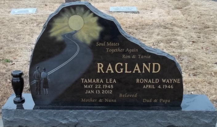Black granite gravestone with engraved mountain road, “RAGLAND,” and floral emblem in a cemetery