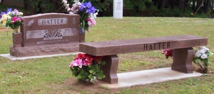 Two engraved memorial benches on grass with pink flowers, one long wooden bench in front and a matching stone seat behind.